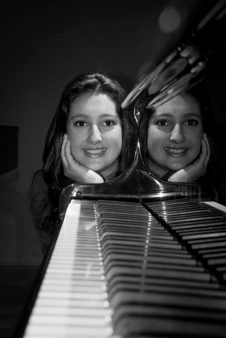 Retrato em preto e branco de Raquel Gonçalves sorrindo, com as mãos sob o queixo; o reflexo do rosto aparece no tampo do piano, acima das teclas.