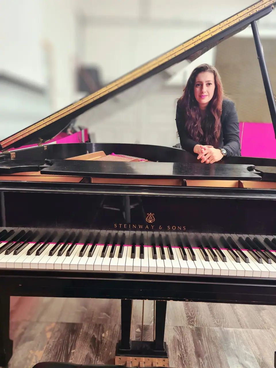 Retrato de Raquel Gonçalves atrás de um piano de cauda aberto, de mãos juntas, olhando para a câmera.